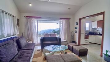 Una sala de estar con sofá, reposapiés y una mesa de centro de vidrio, un balcón con cortinas, y una cocina al fondo.