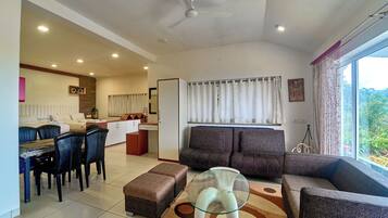 A modern living room with a dining area, a sofa set, a glass coffee table, and a view of greenery outside.