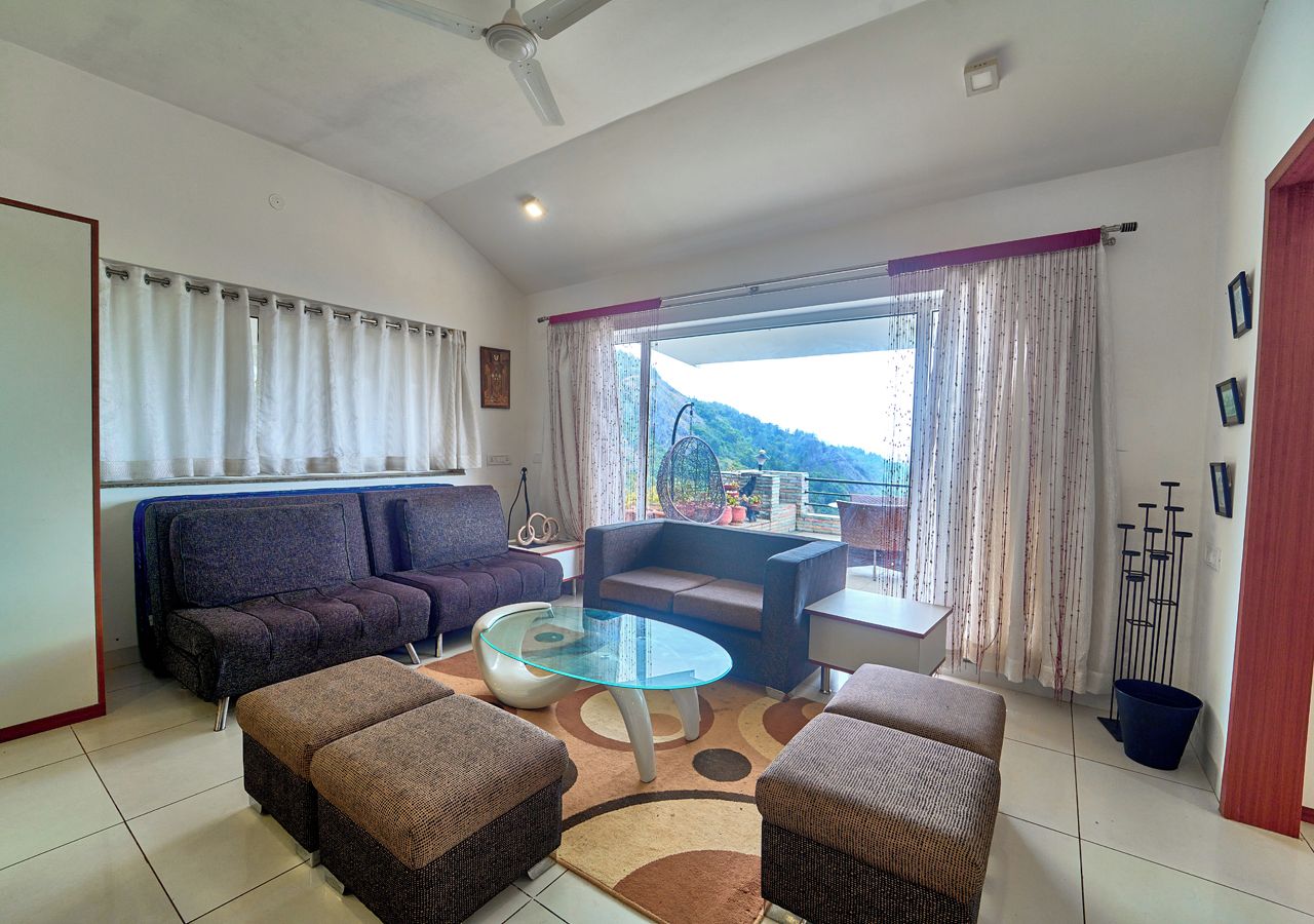 A living room with a sofa, ottomans, a glass coffee table, and a view of a mountainous landscape through the window.