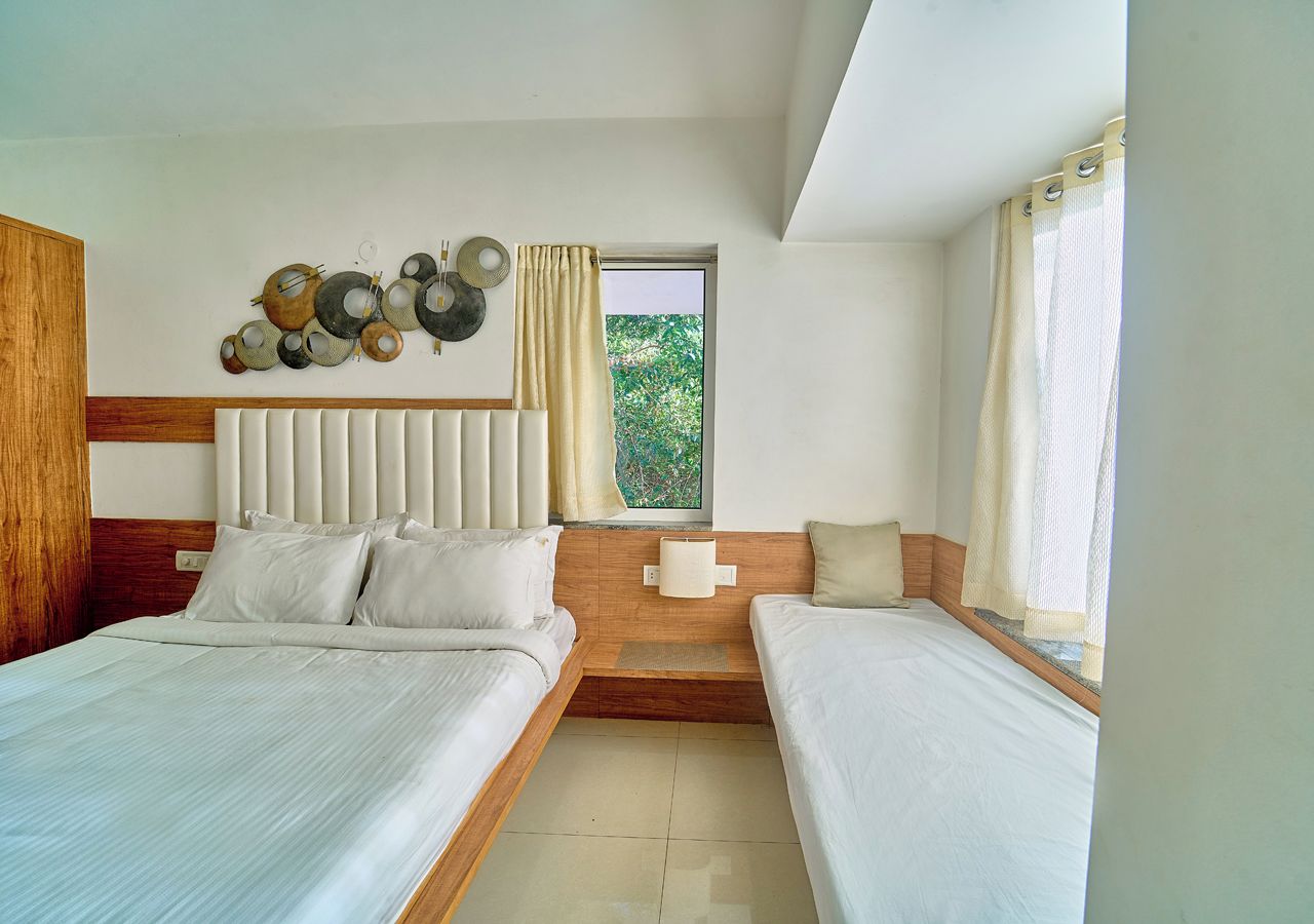 A hotel room with two beds, a wooden headboard, a wall art piece, a window with curtains, and a view of greenery outside.