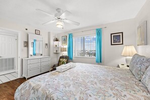 Un dormitorio con cama, ventilador de techo, ventana con cortinas, mesita de noche con lámpara y cómoda.