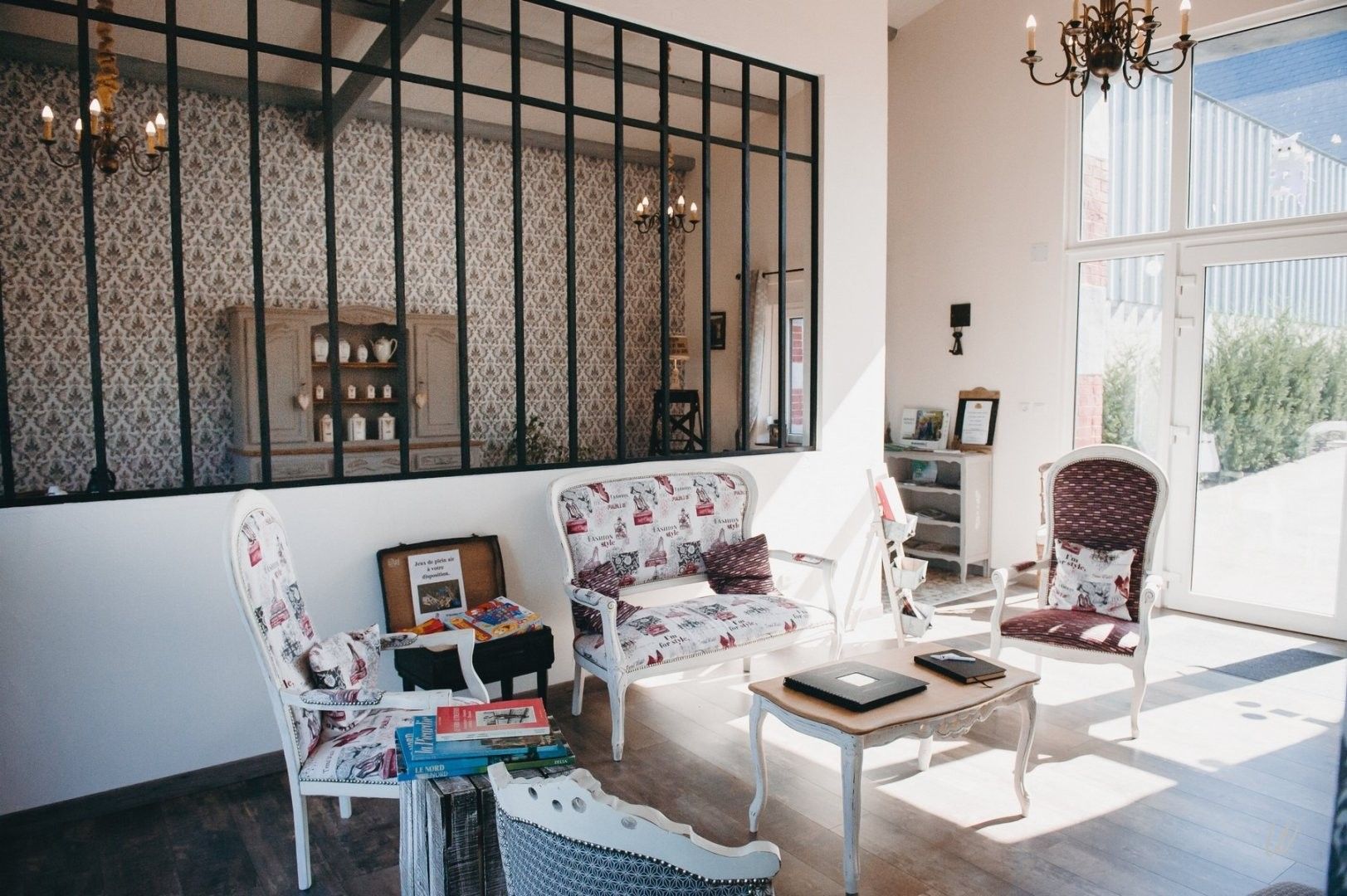 Une pièce bien éclairée, dotée d’une grande fenêtre, de deux fauteuils rembourrés, d’une petite table avec des livres et d’un élément décoratif mural.
