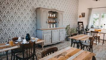Un coin repas avec des tables et des chaises en bois, un buffet ancien contenant de l’ustensile de cuisine, un papier peint à motifs et un lustre.