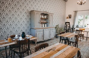 Et spiseområde med træborde og stole, et antikt skab med køkkenudstyr, tapet med mønster og en lysekrone.