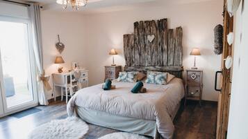 Une chambre à coucher avec un grand lit, deux tables de chevet, un bureau et une chaise. On y trouve une grande fenêtre avec des rideaux, un lustre et une décoration murale de style rustique.