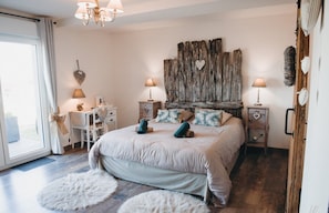 A bedroom with a large bed, two bedside tables, a desk, and a chair. There is a large window with curtains, a chandelier, and a rustic wall decoration. - Bed and Breakfast 'Chambres Azélice' with Shared Terrace, Shared Garden and Wi-Fi (Féron)