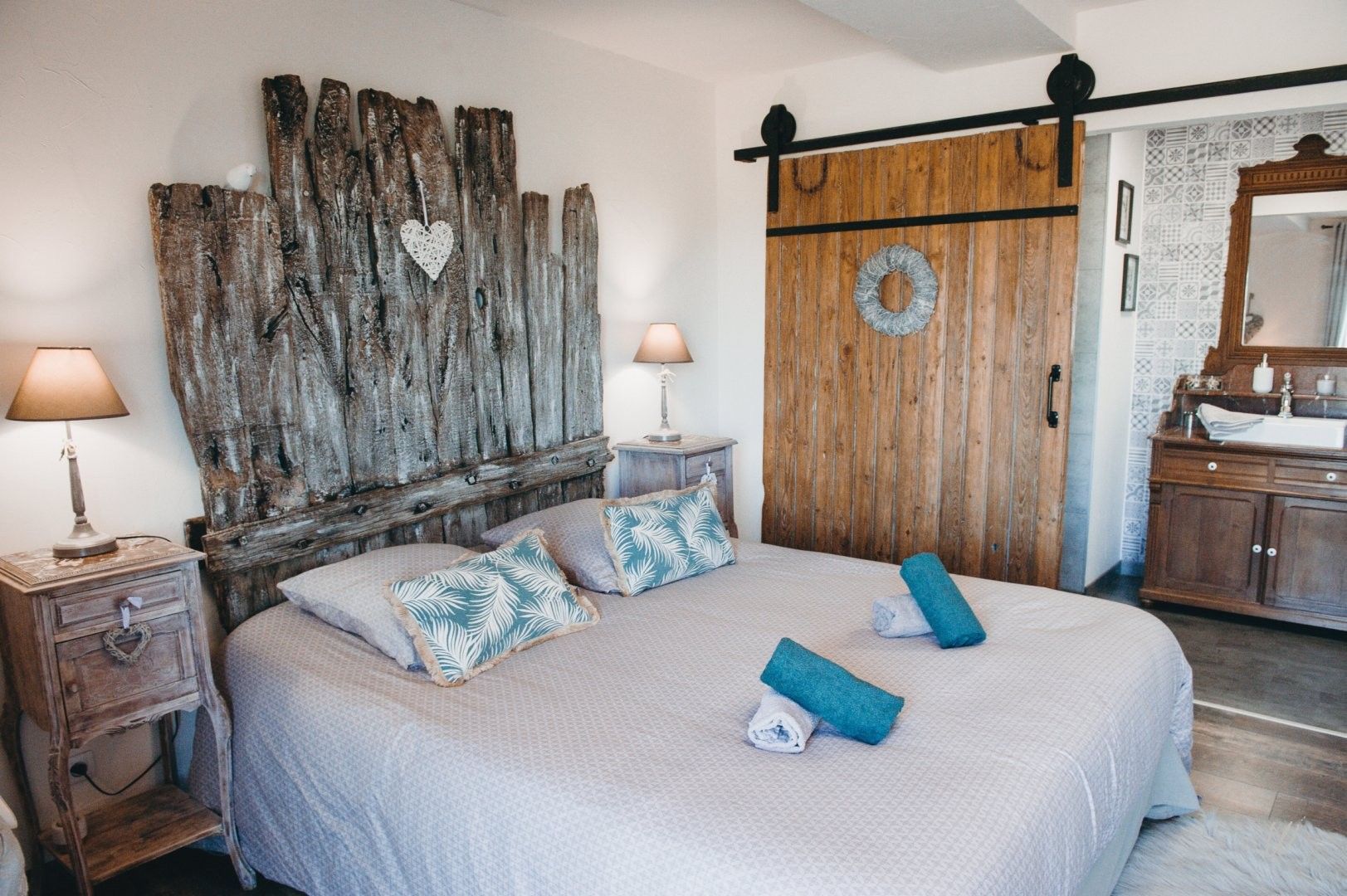 Een slaapkamer met een rustiek houten hoofdeinde, een bed met gekleurde kussens, een nachtkastje met een lamp en een garderobe met een spiegel.