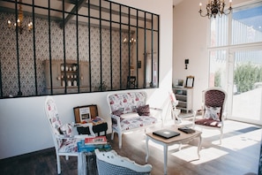 A well-lit room with a large window, two upholstered chairs, a small table with books, and a decorative wall piece. - Bed and Breakfast 'Chambres Azélice' with Shared Terrace, Shared Garden and Wi-Fi (Féron)