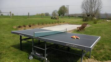 Une table de ping-pong portable sur roulettes, installée sur un terrain herbeux, avec un petit espace pour s’asseoir et des arbres en arrière-plan.