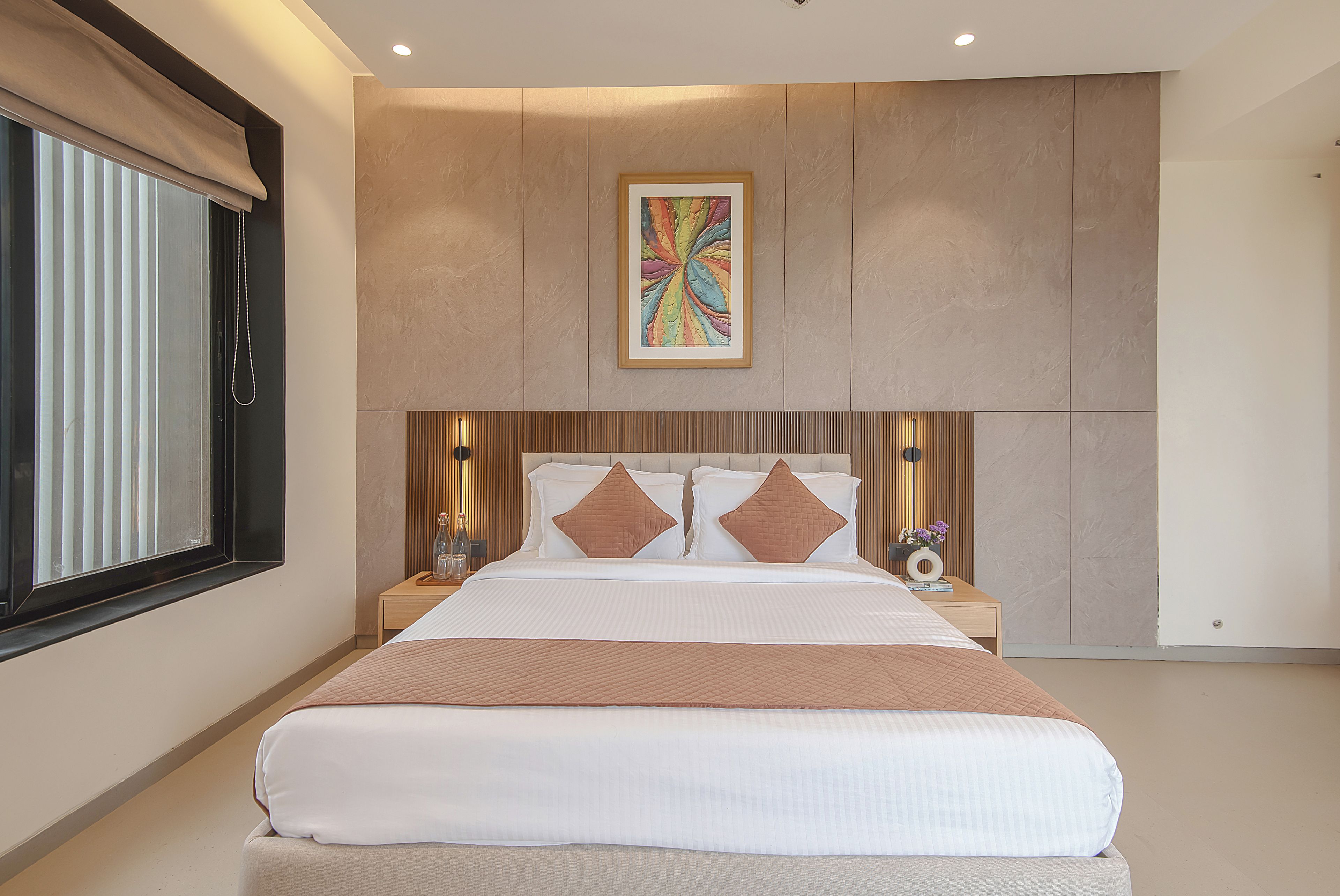A modern bedroom with a large bed, a headboard with a built-in artwork, bedside tables with lamps, and a window with blinds.