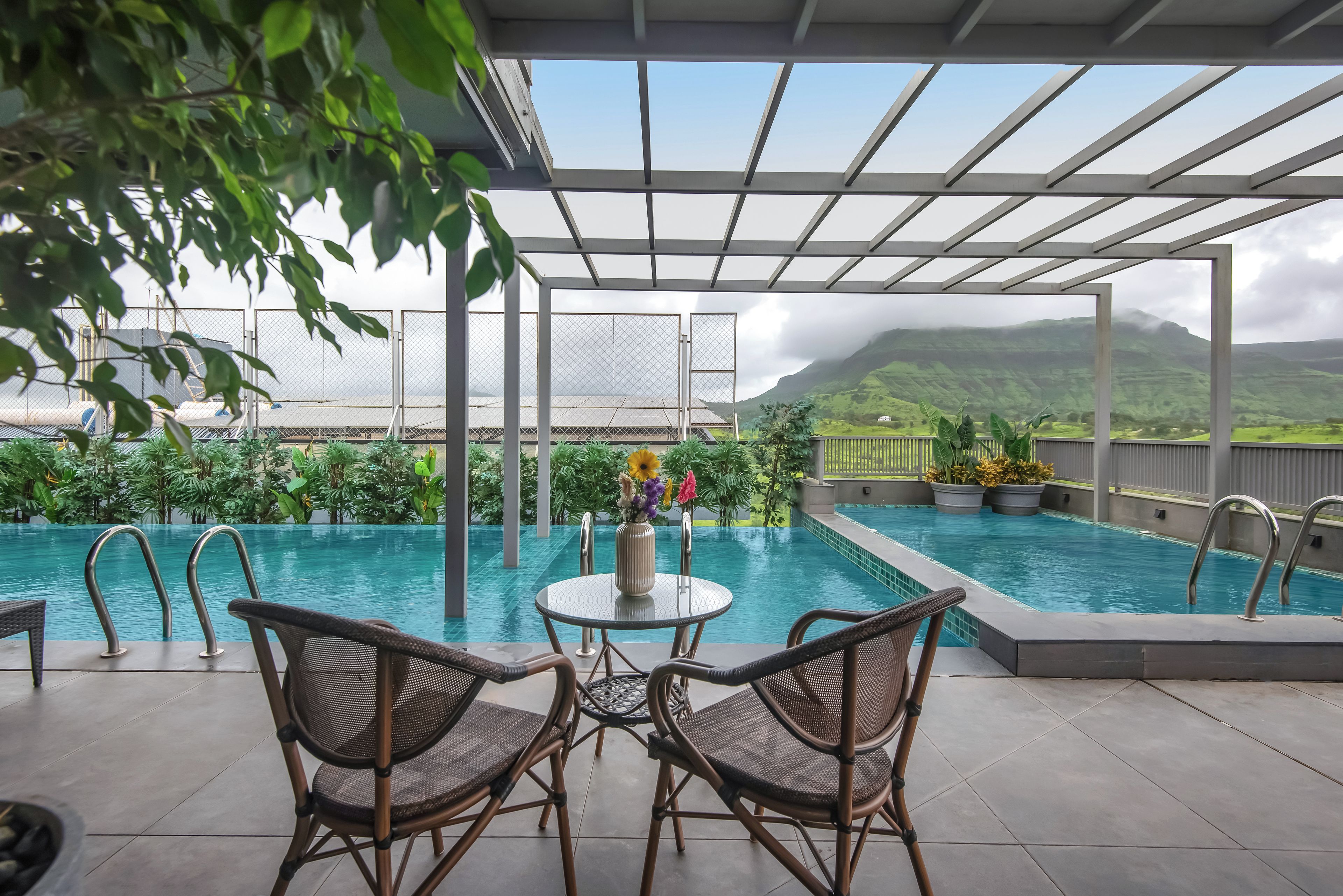 An outdoor pool area with two wicker chairs, a table with a vase of flowers, and a view of a mountainous landscape.