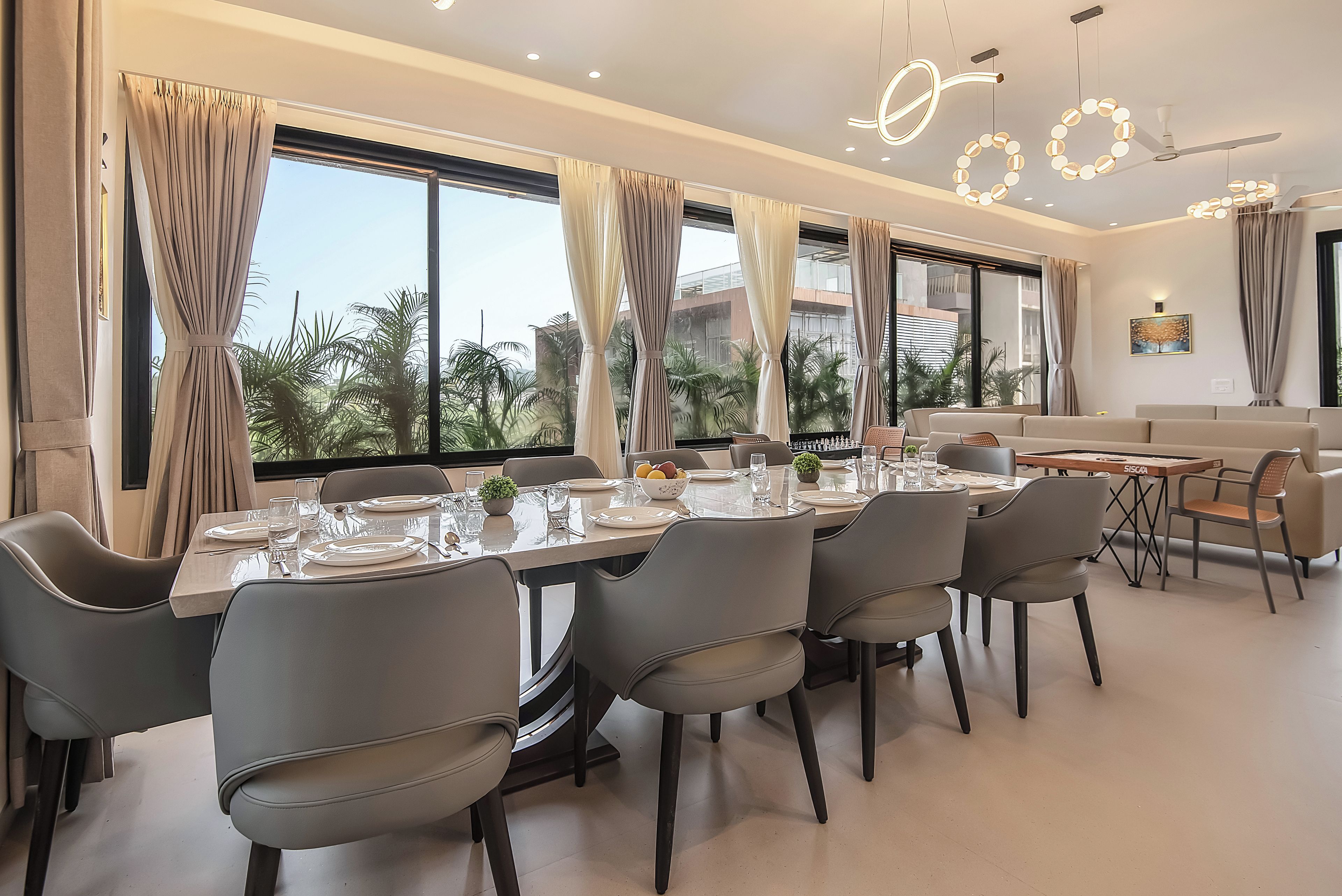 A modern dining area with a long table, multiple chairs, and a view of palm trees through large windows.