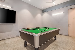 Una mesa de billar con bolas y tacos, un televisor montado en la pared, y una puerta que dice 'KITCHEN' en una habitación con paredes con textura.