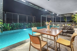 Un área de piscina al aire libre con una mesa y sillas, rodeada de plantas y una cerca.