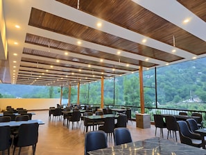 Un amplio espacio interior con mesas y sillas, ventanas grandes que ofrecen vistas a un paisaje montañoso boscoso, y un techo de madera con iluminación empotrada.