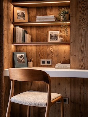 A wooden bookshelf with books and framed pictures, a desk with a chair, and a power outlet. - Chalet Mara (Les Houches)
