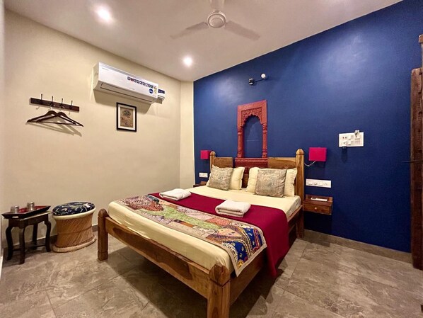 A bedroom with a wooden bed, a red and white bedspread, a ceiling fan, an air conditioning unit, a small table with a basket, and a wall-mounted shelf with a picture.