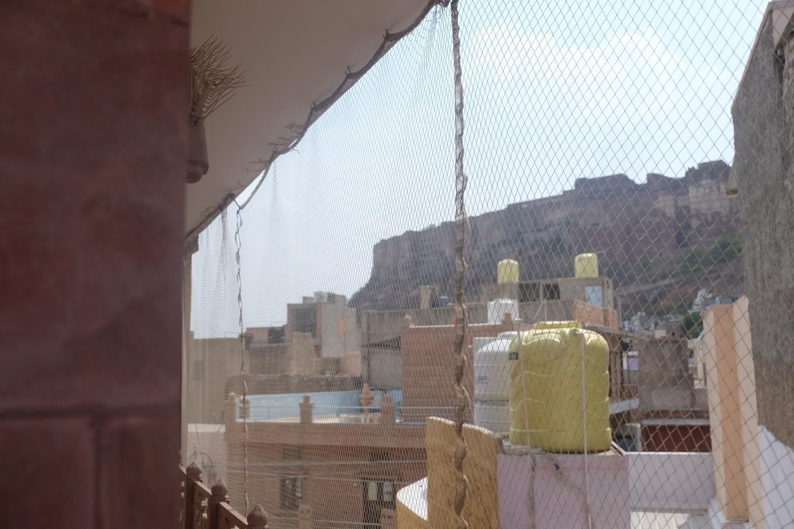 Vista a través de una ventana con una cerca de malla, que da a una zona residencial con edificios y una colina al fondo.