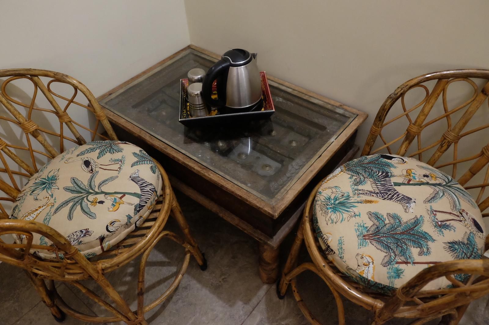 Una mesa pequeña con una tetera y una bandeja de condimentos, junto a dos sillas de mimbre con cojines estampados.