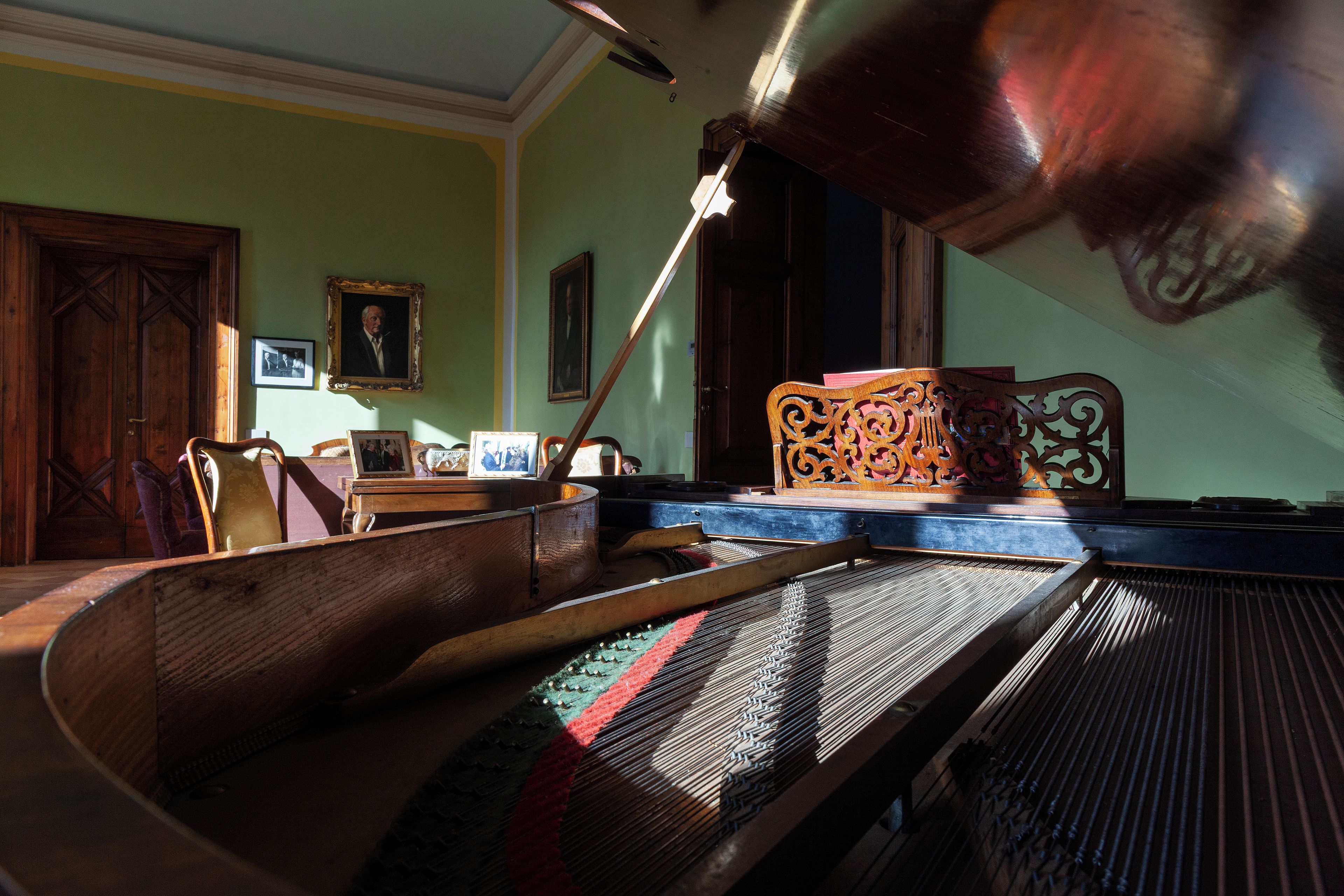 Una habitación con un piano, una silla de madera, una mesa con una lámpara y cuadros enmarcados en la pared.