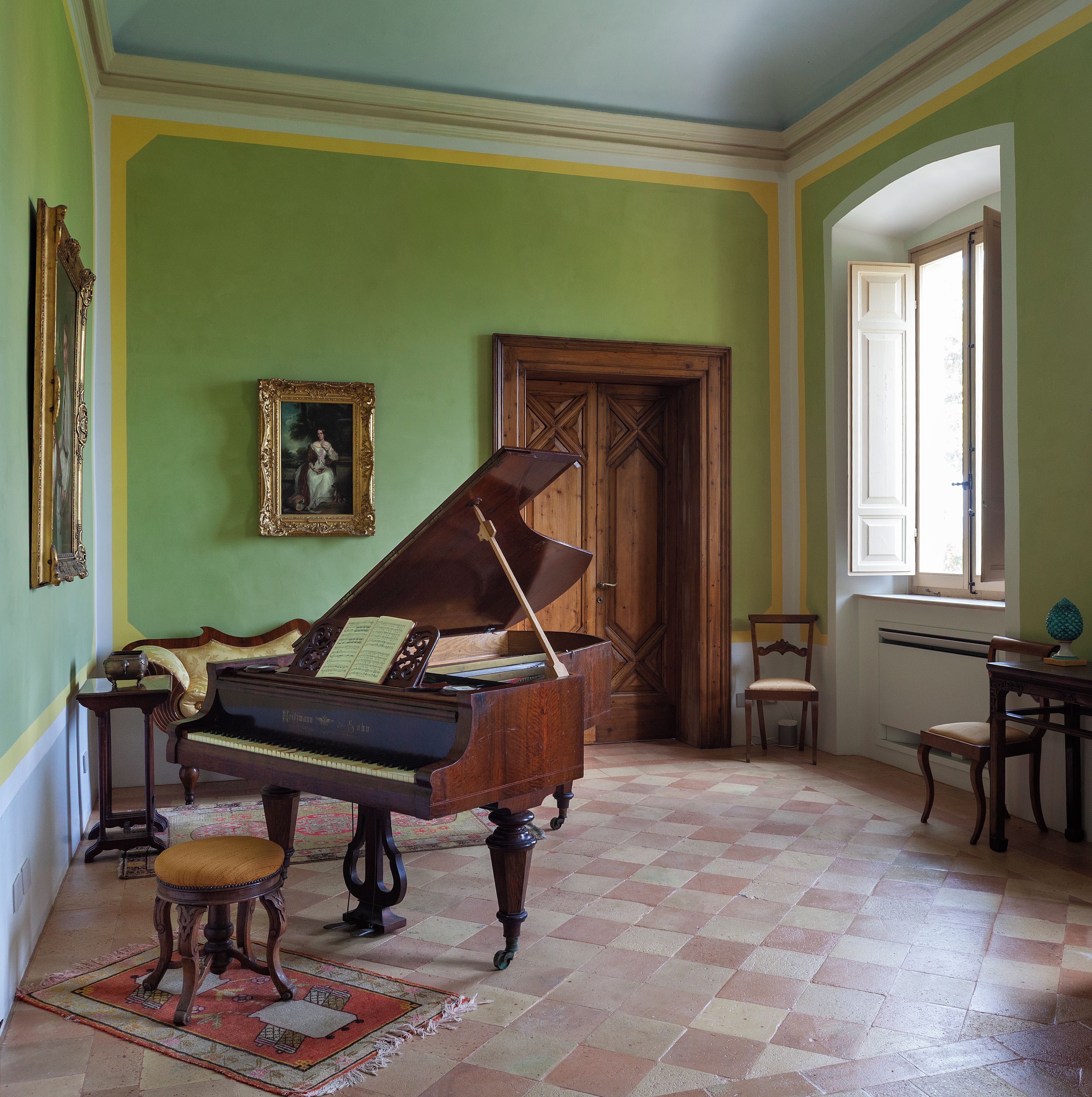 Um quarto com um piano de cauda, uma porta de madeira, um quadro emoldurado e uma janela.