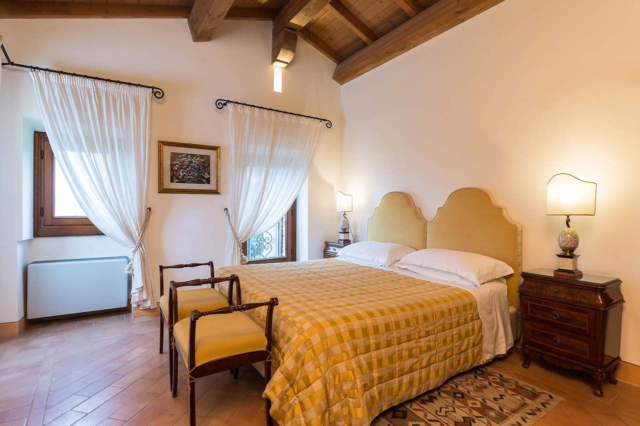 Quarto com cama, duas cadeiras, mesa de cabeceira, abajur e janela com cortinas.