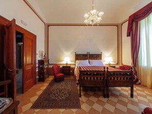 Habitación de hotel con piso a cuadros, una cama con colcha a rayas, un sillón tapizado rojo, una mesita de noche de madera, una lámpara, un candelabro y una puerta con un cartel.