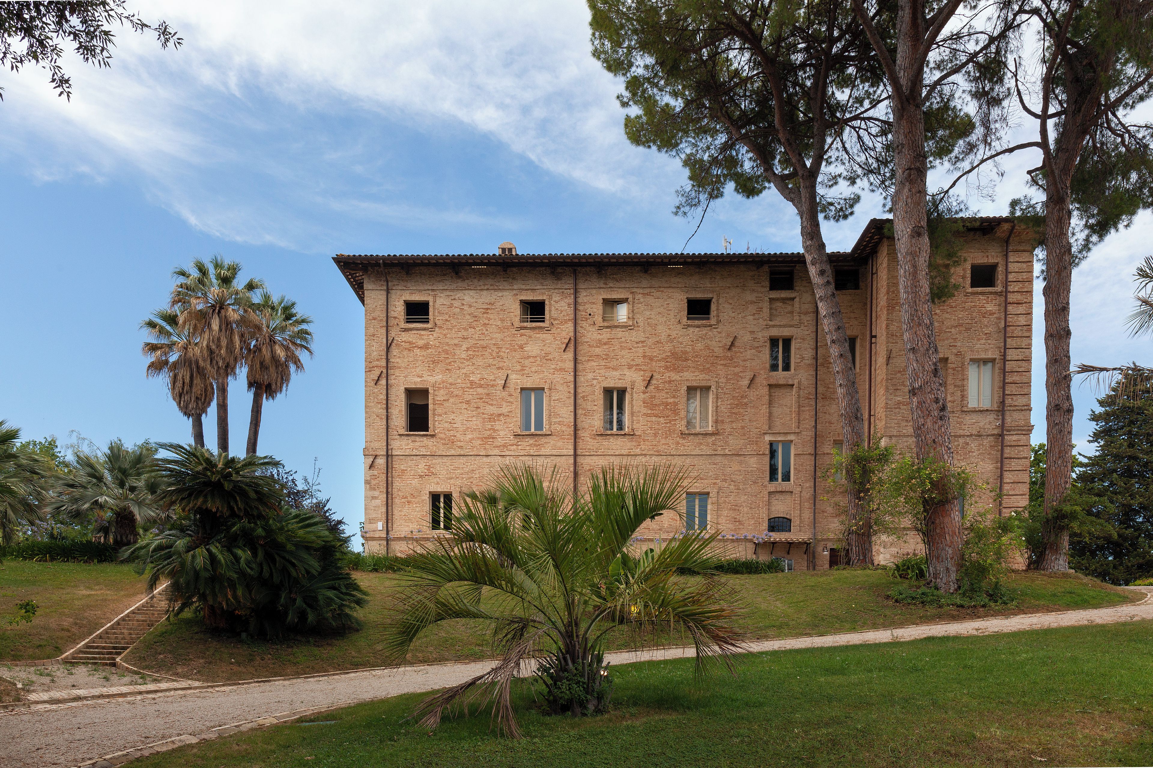 Un immeuble à plusieurs étages, doté d’une façade en briques, de nombreuses fenêtres et entouré d’un paysage agrémenté de palmiers et d’un chemin de gravier.