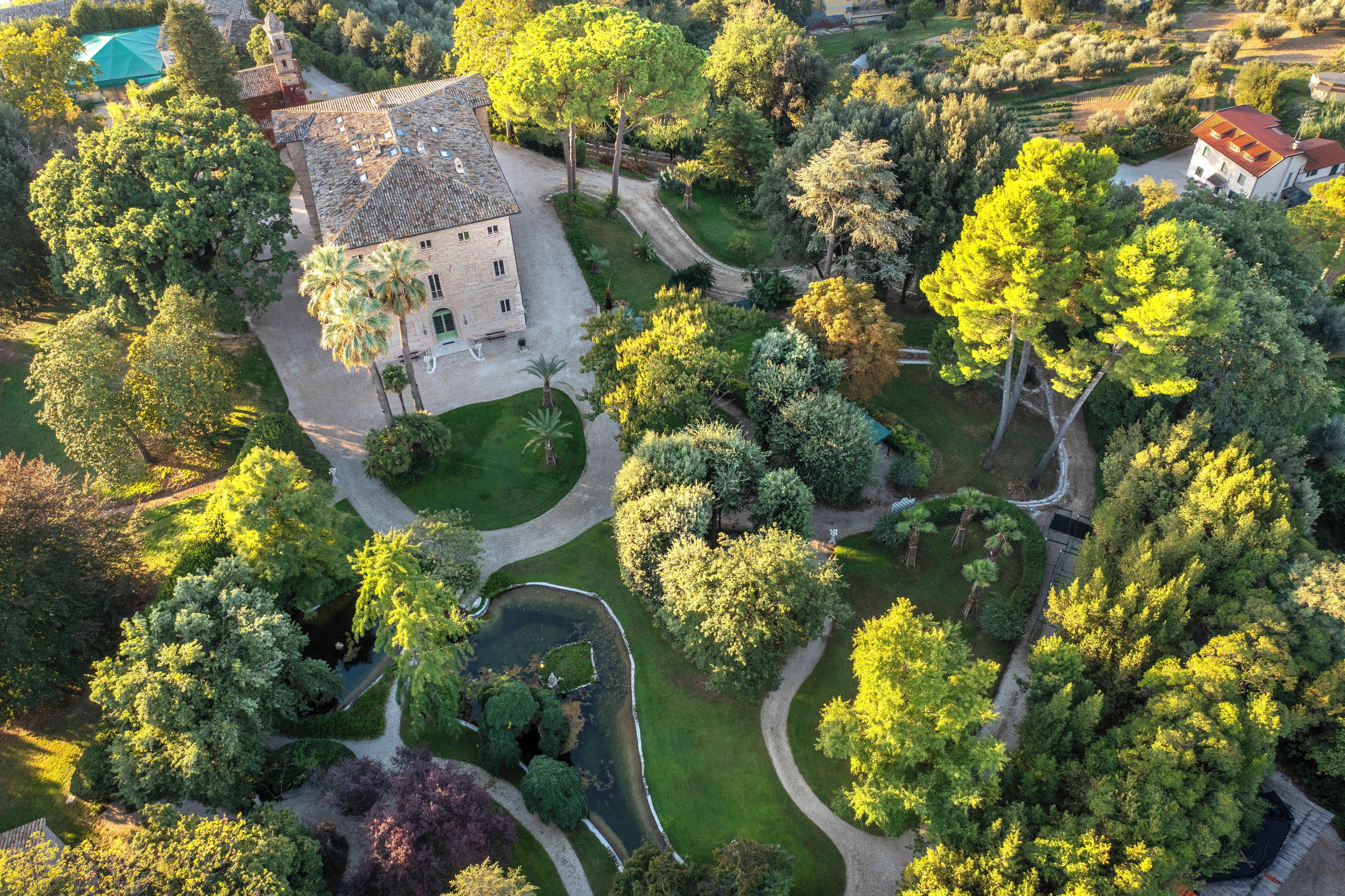 An aerial view of a property with a central building surrounded by lush greenery, a pond, and a winding path.