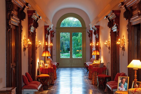 A hallway with a central door leading to a garden, flanked by ornate columns and lit by wall sconces.