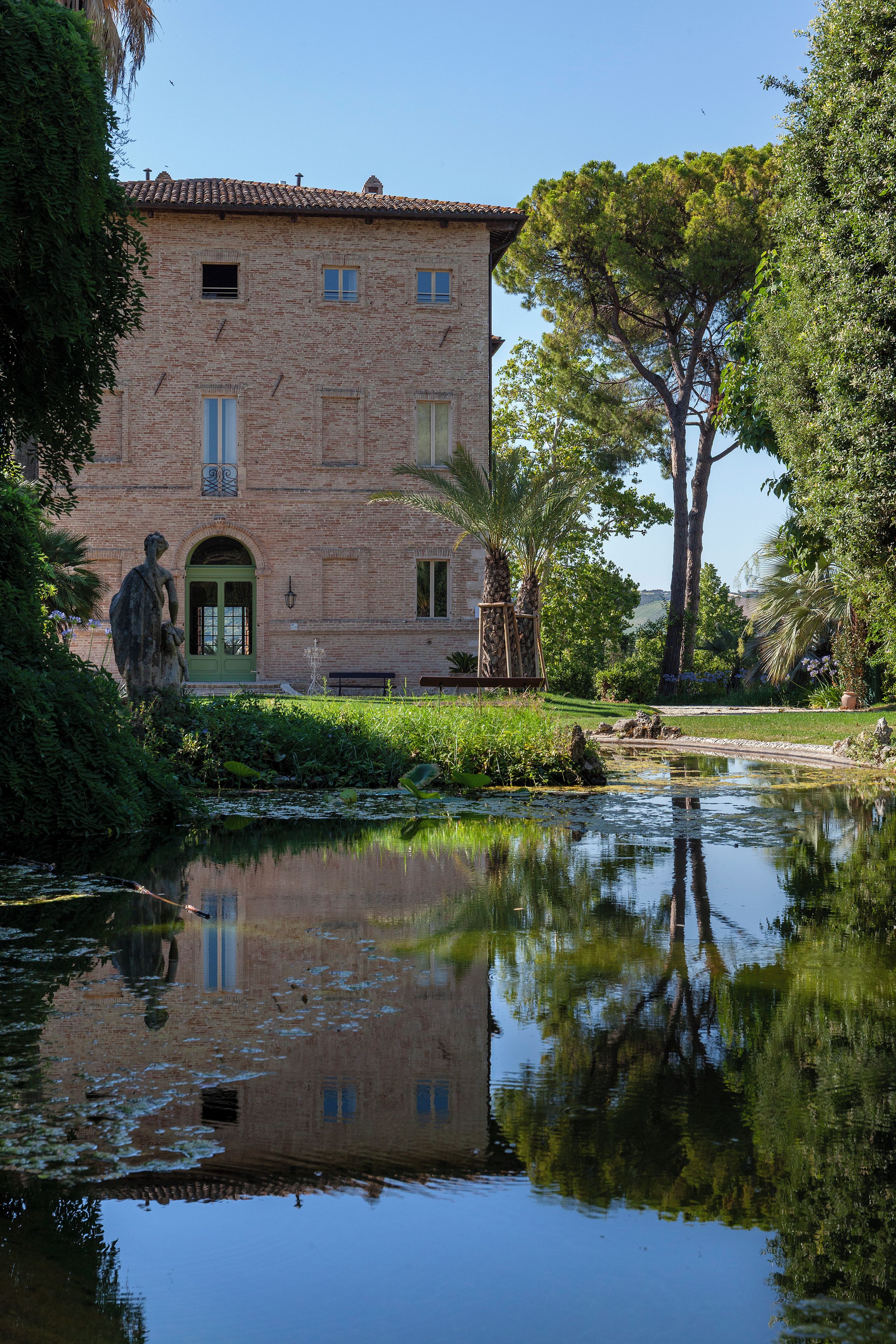 Un edificio histórico con su reflejo en aguas tranquilas, rodeado de follaje y un cielo despejado.