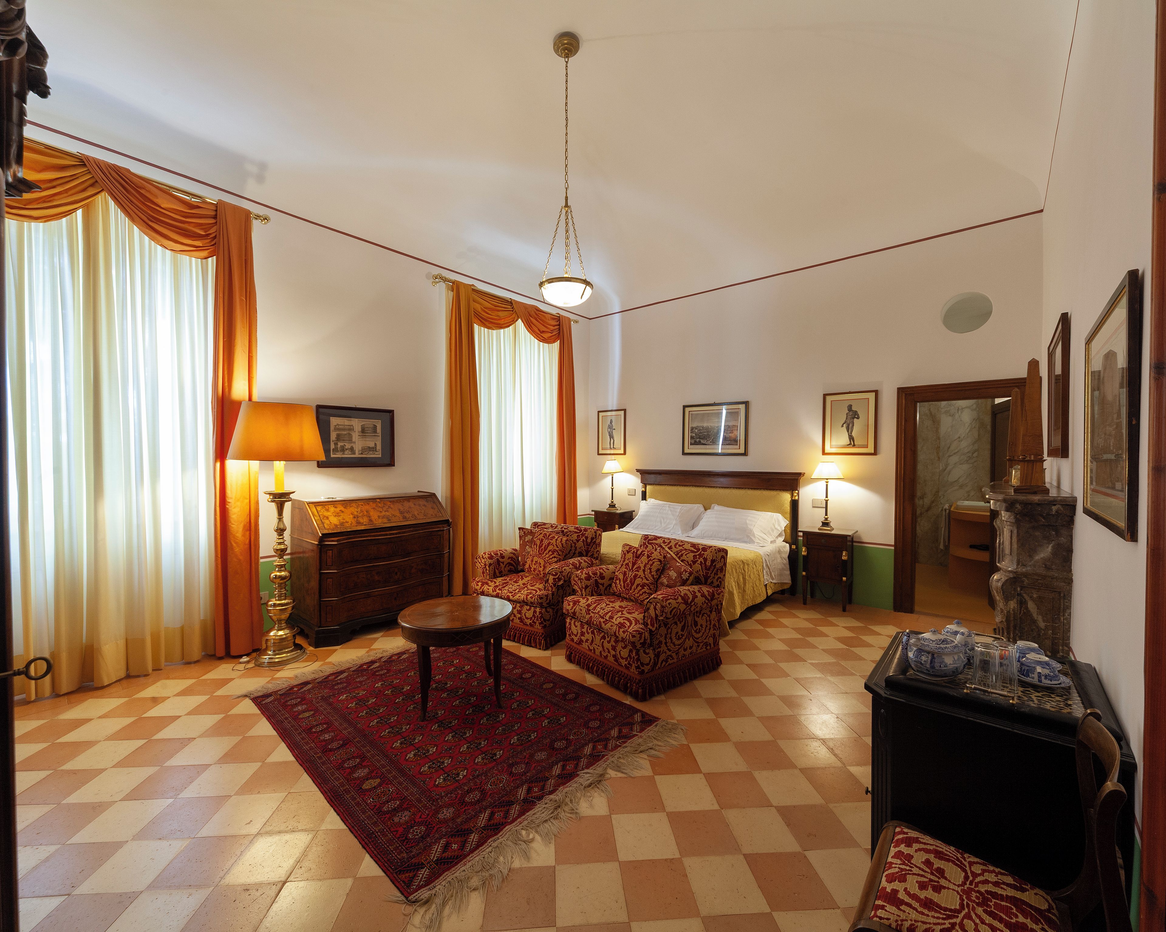A hotel room with a bed, two armchairs, a small table, a lamp, a rug, and a fireplace.