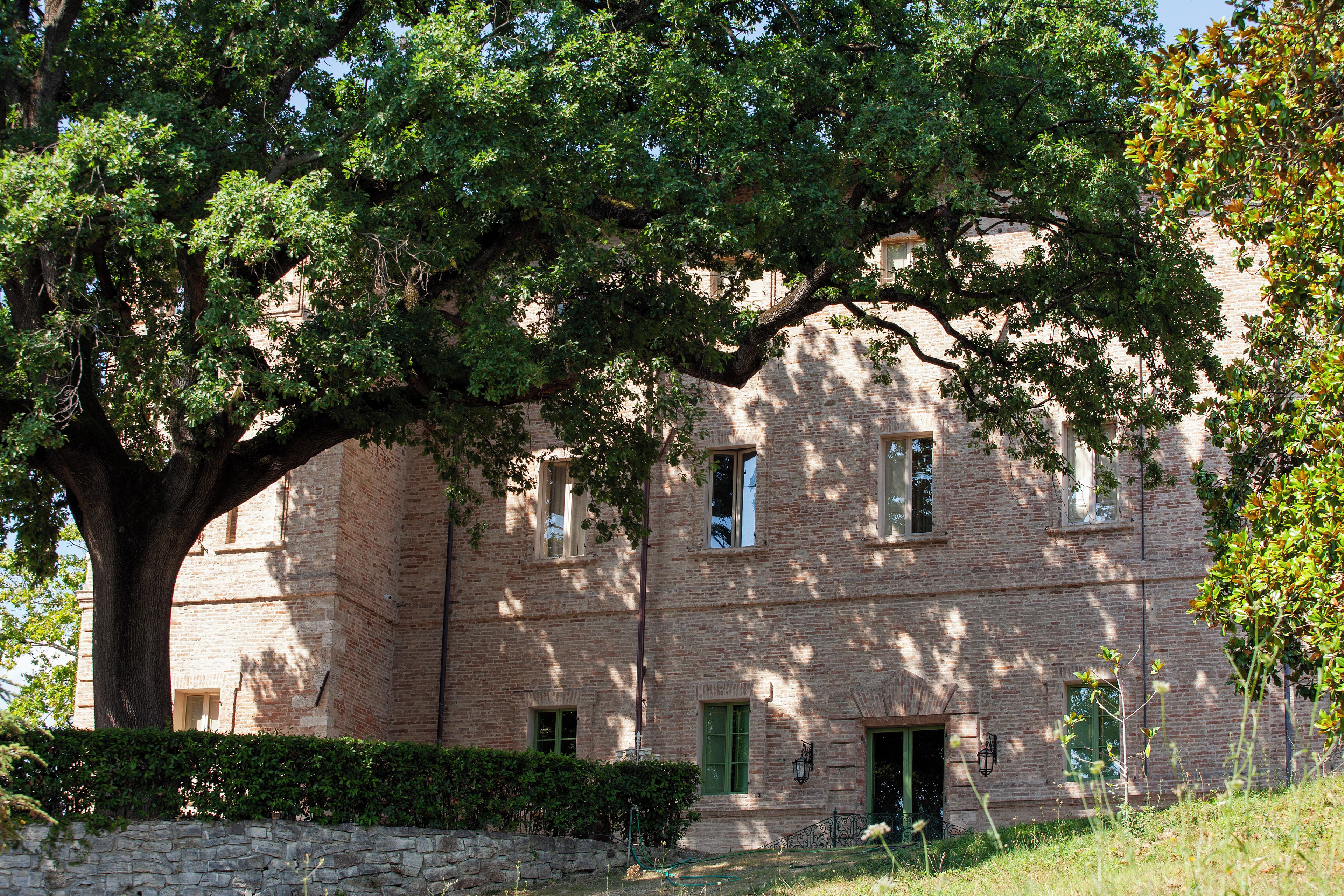 A brick building with a large tree in front, a stone wall, and a grassy area.