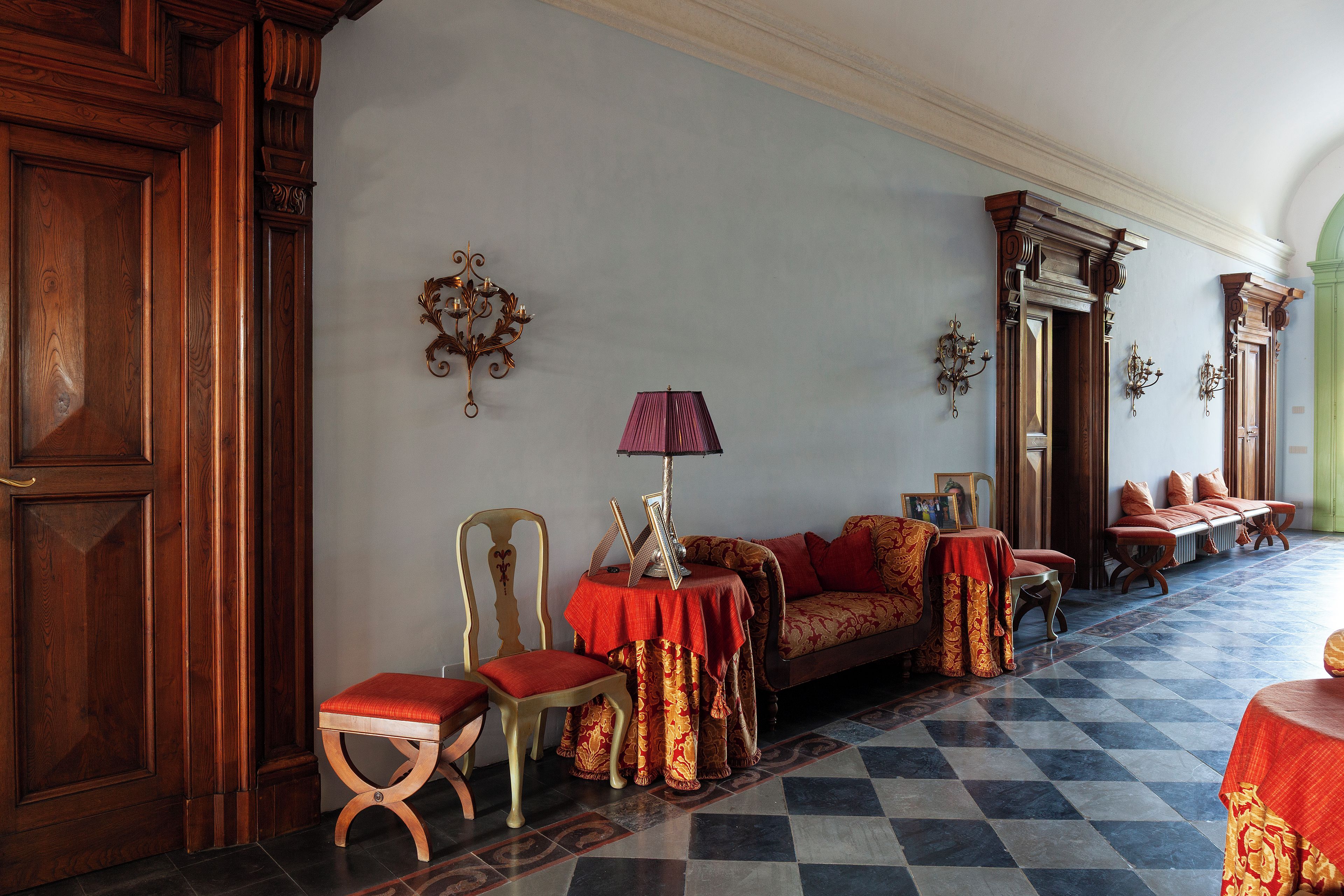 A room with a checkered floor, a sofa with red and gold upholstery, a wooden chair, a table with a lamp, and wall-mounted sconces.