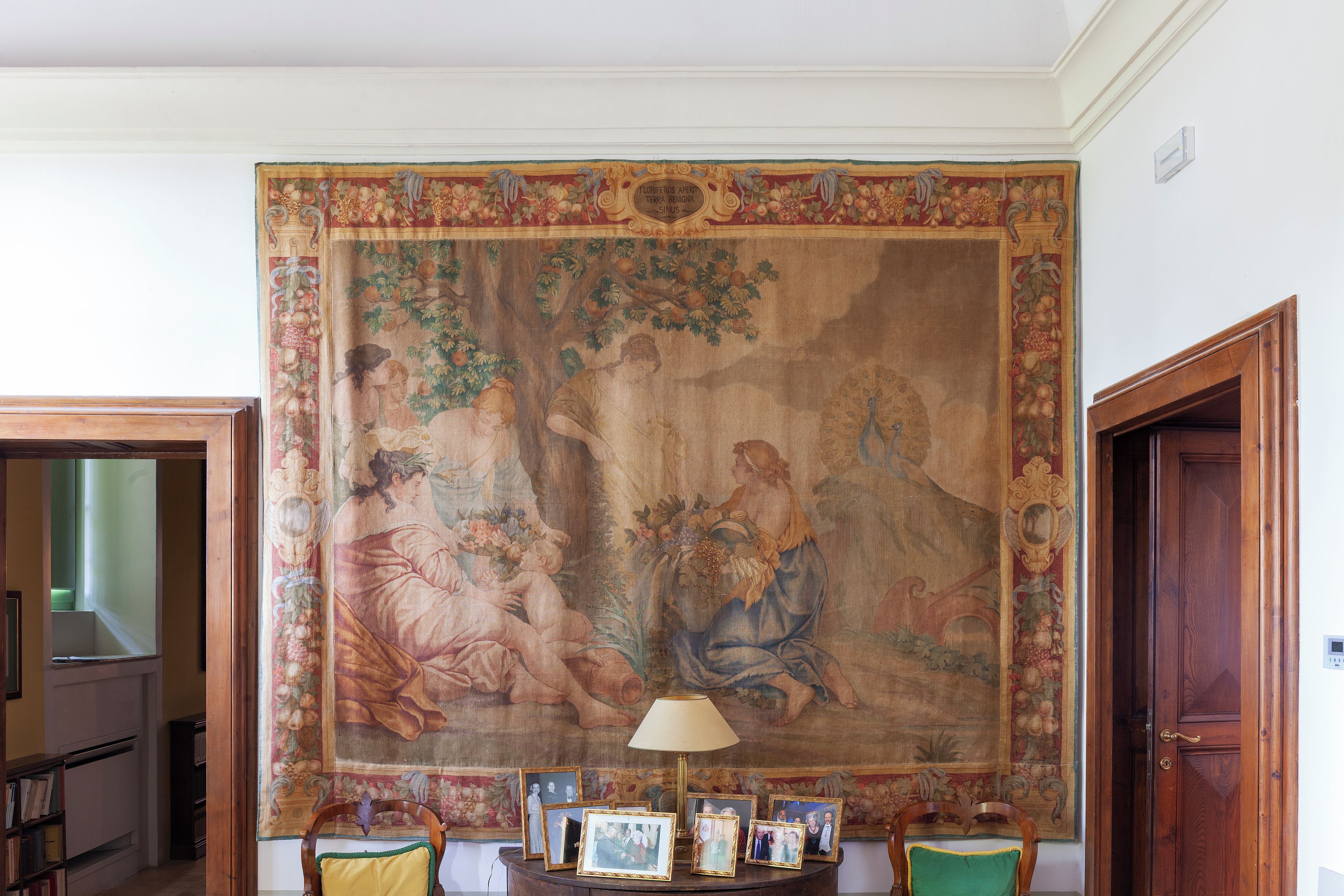 A room with a large tapestry depicting a pastoral scene, a wooden door, and a table with framed photographs.