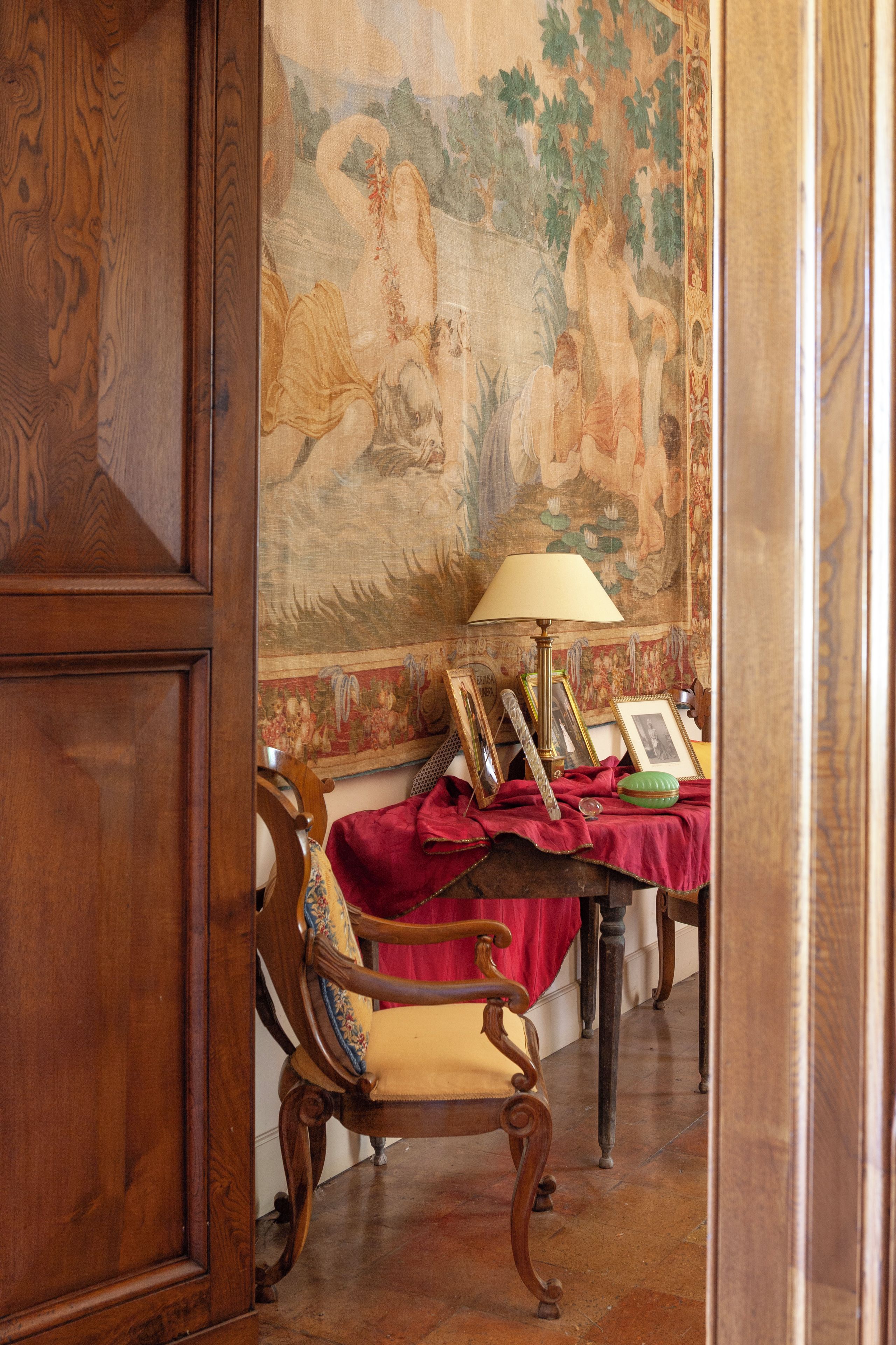 A room with a wooden chair, a table with a lamp, and a tapestry on the wall.