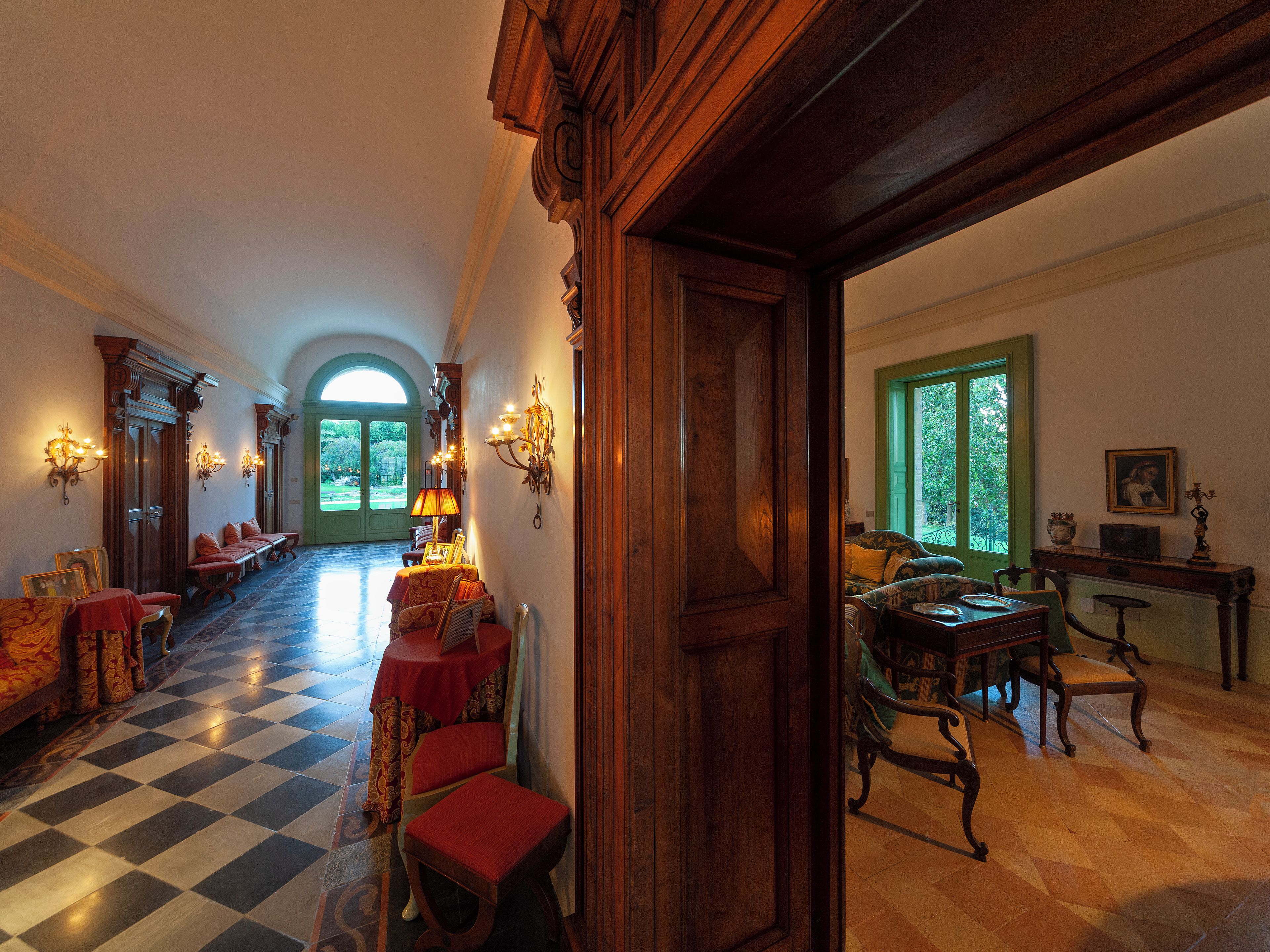 A spacious hallway with a checkered floor, wooden furniture, and a view of a garden through a green door.