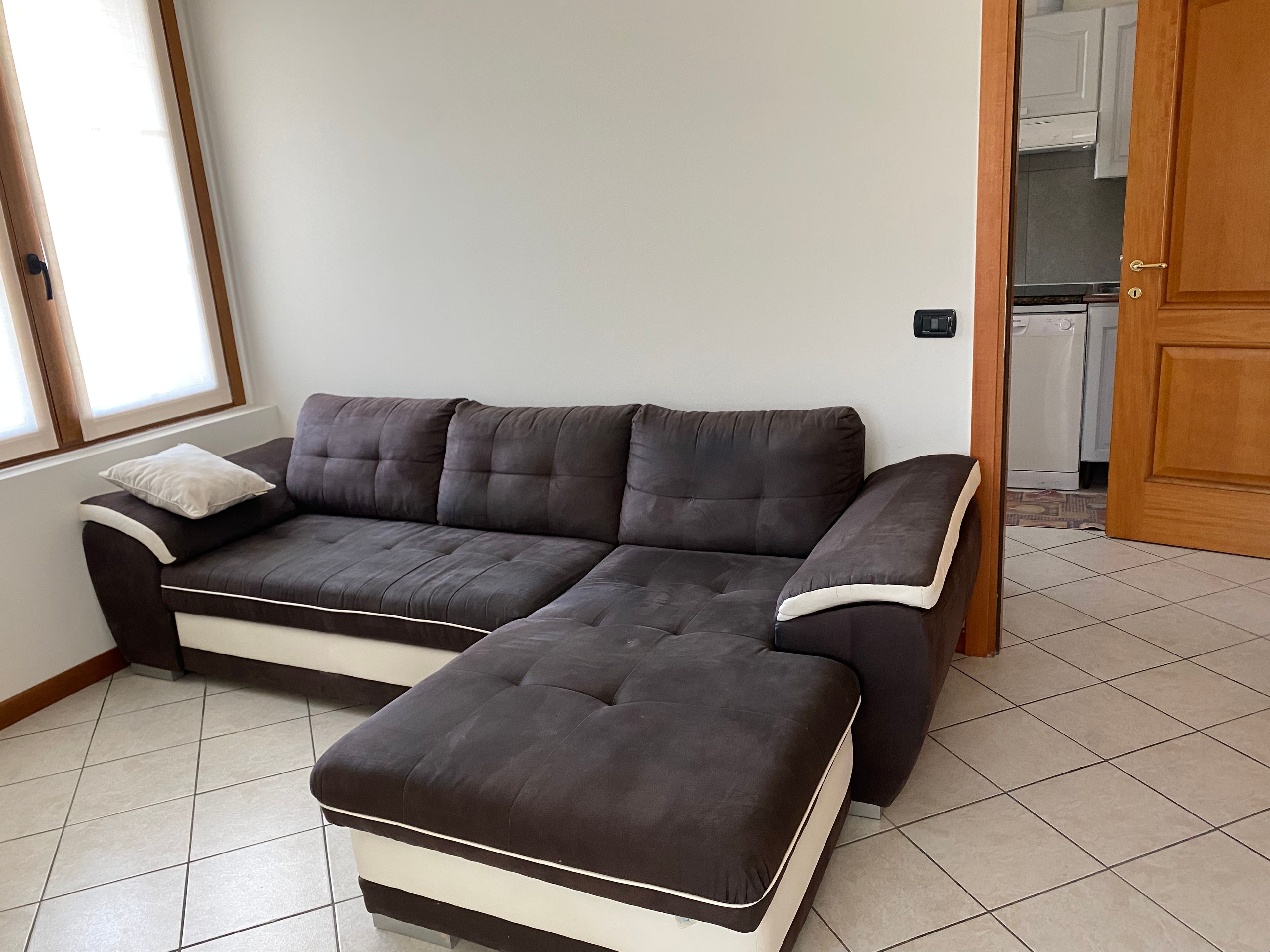 Un sofá modular con un reposapiés a juego en una habitación con suelo de baldosas y una cocina visible.
