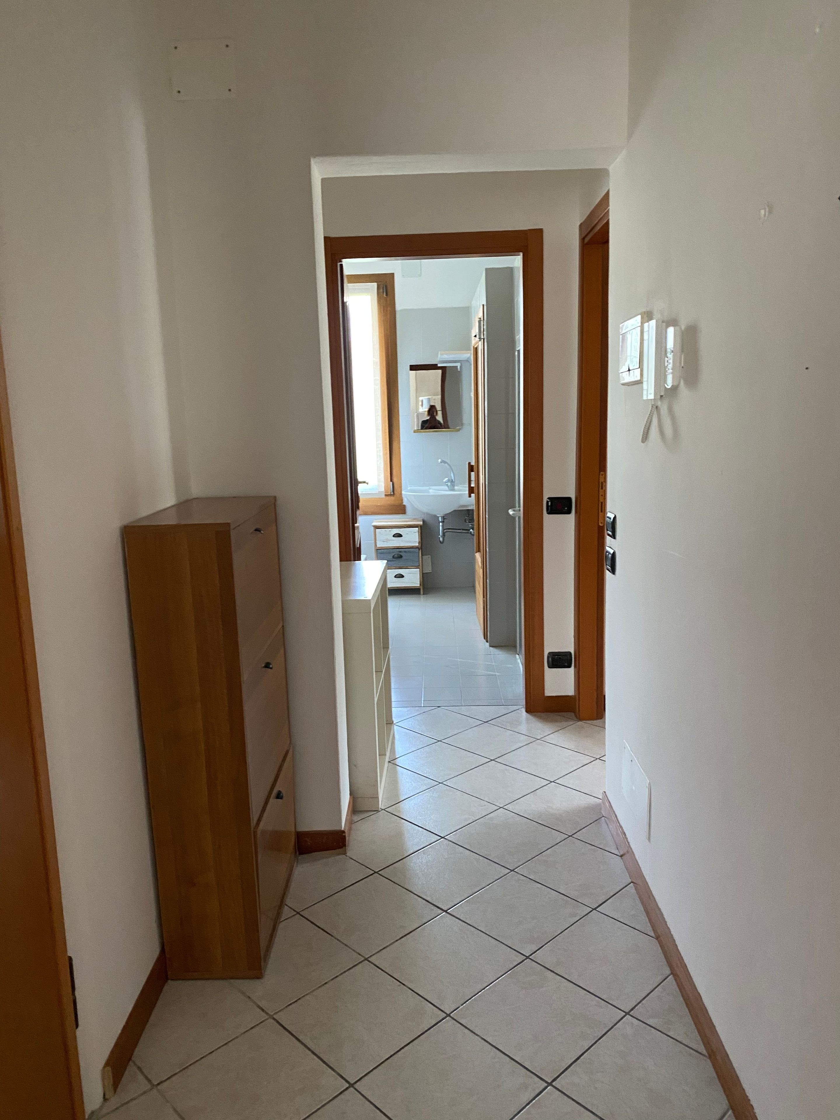 A narrow hallway with a wooden cabinet, a tiled floor, and an open door leading to a bathroom with a sink and mirror.
