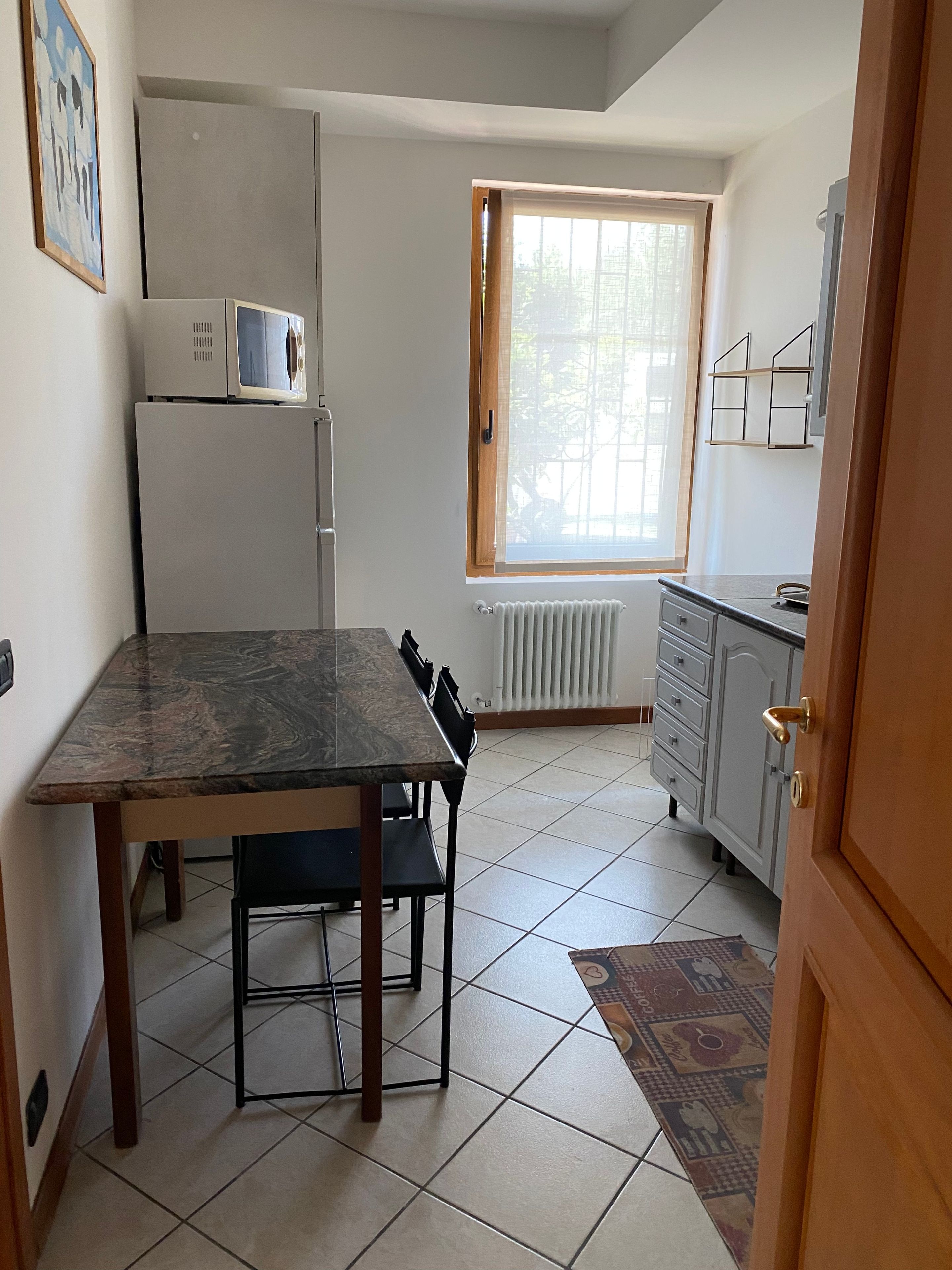 Una cocina compacta con mesa, sillas, refrigerador, microondas, ventana con vistas y radiador.