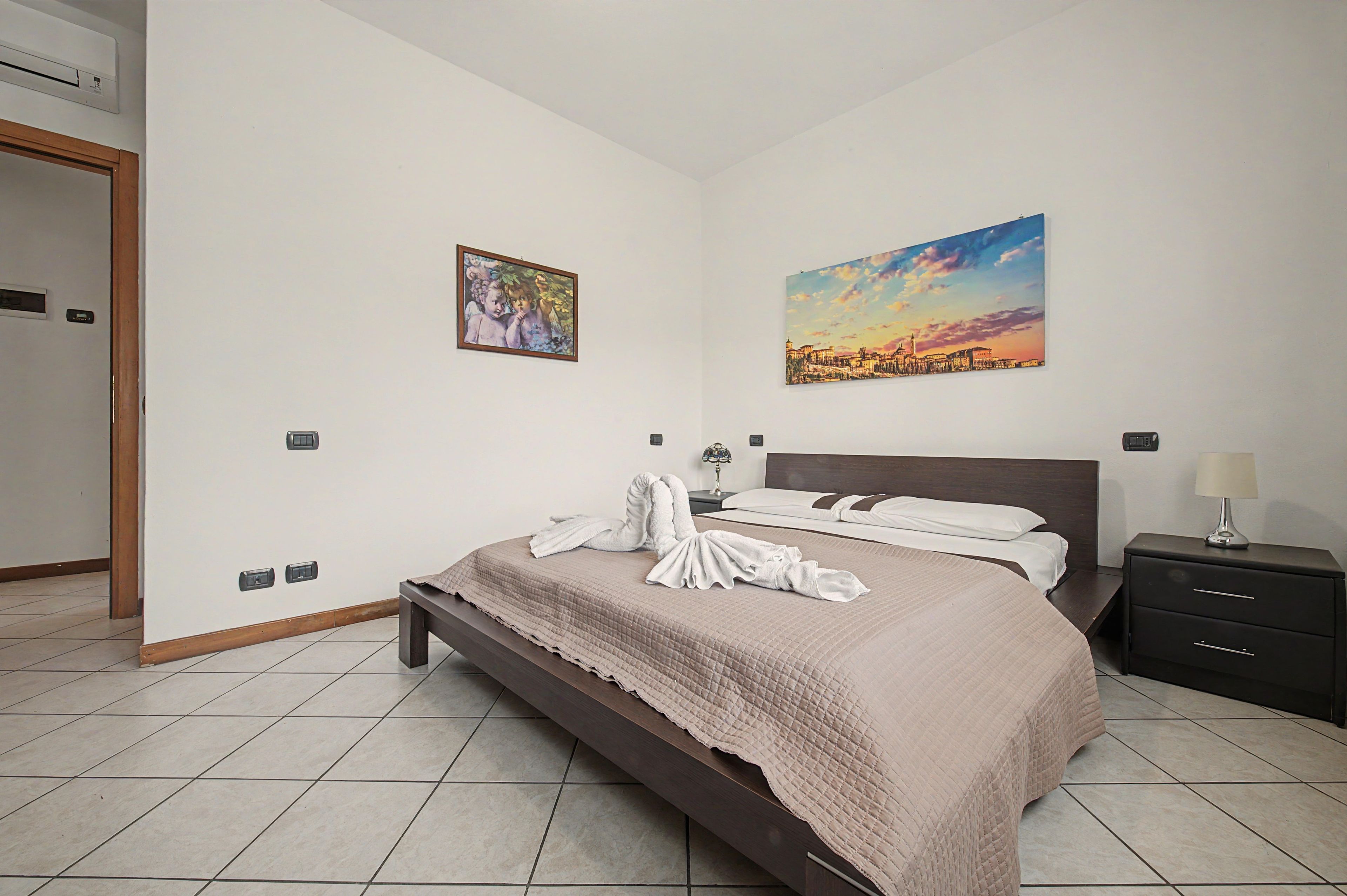 Habitación de hotel con cama, mesita de noche, lámpara, un cuadro y aire acondicionado instalado en la pared.