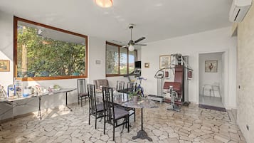 Ein Zimmer mit Esstisch, Stühlen, Deckenventilator, Trainingsgeräten und einem großen Fenster mit Blick auf Bäume.