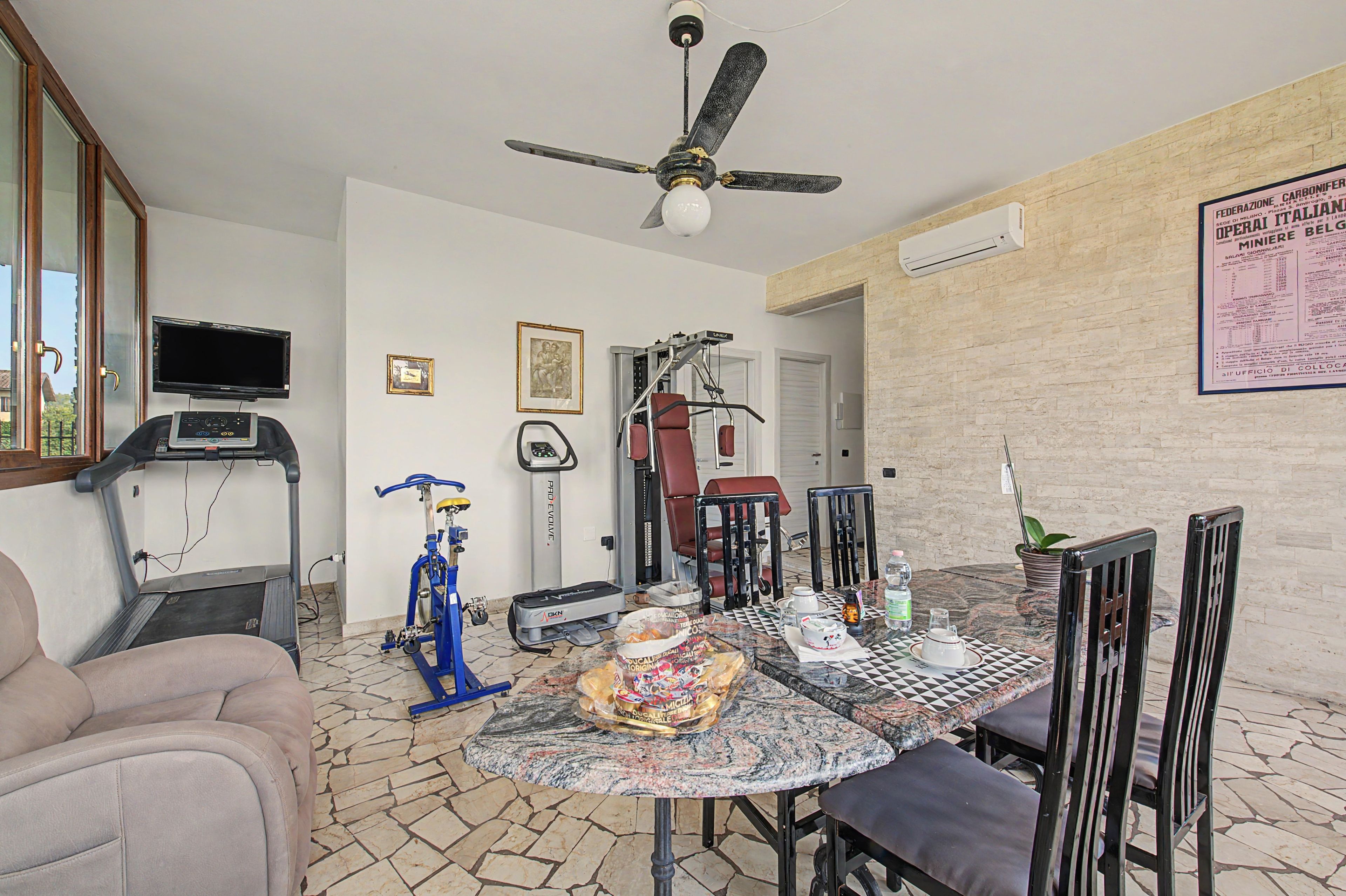A room with a treadmill, stationary bike, exercise equipment, a ceiling fan, a dining table with chairs, and a wall with a menu.