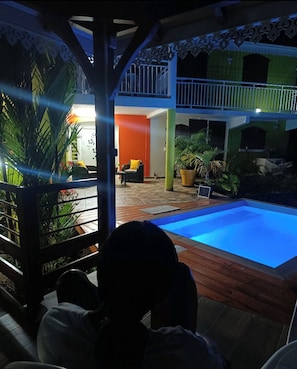 A pool area with a wooden deck, a covered seating area, and a view of a building with a balcony. - T -SYSSY LODGE  FOR 2 ADULTS (WITHOUT CHILDREN) TO DISCOVER MARTINIQUE (Fort-de-France)