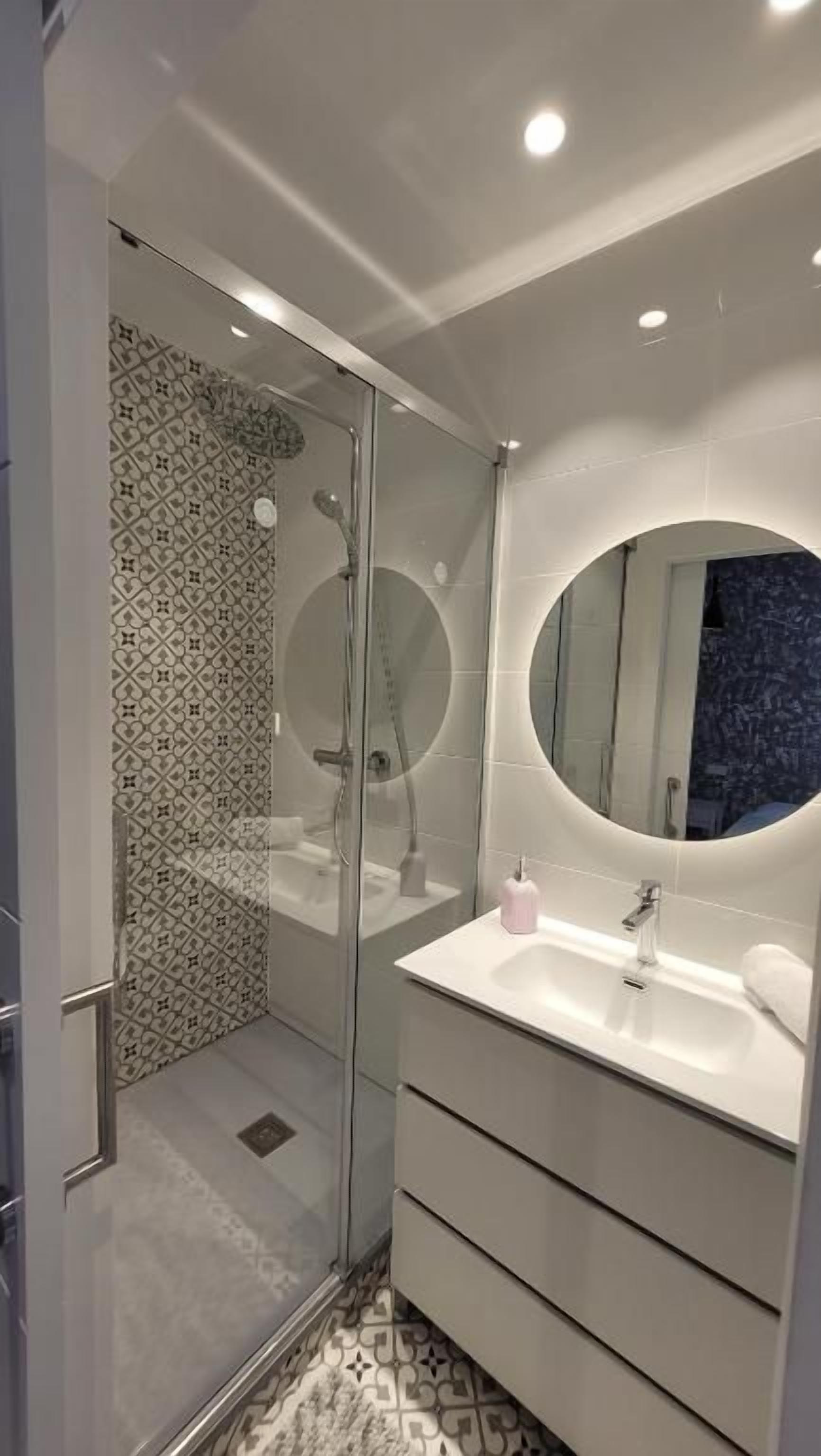 Een moderne badkamer met een douche met glazen wand, een ronde spiegel en een witte wastafel.