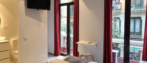A hotel room with a bed, a TV, a desk, and a balcony with red curtains.