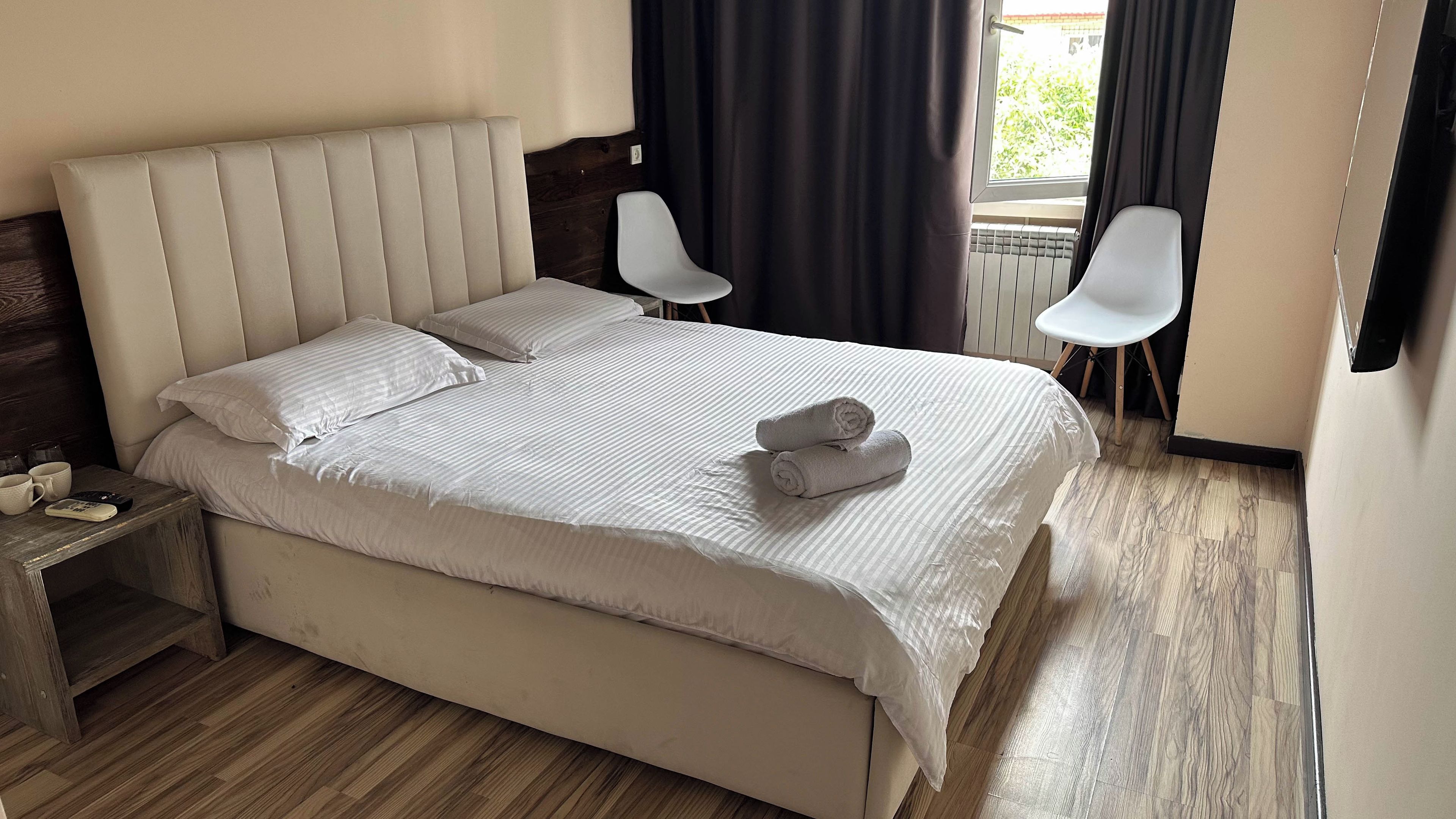 Una cama bien hecha con almohadas y una toalla enrollada, una mesita de noche de madera con un control remoto y una taza, y dos sillas blancas en una habitación con suelo de madera y una ventana con cortinas.