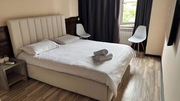 Una cama bien hecha con almohadas y una toalla enrollada, una mesita de noche de madera con un control remoto y una taza, y dos sillas blancas en una habitación con suelo de madera y una ventana con cortinas.