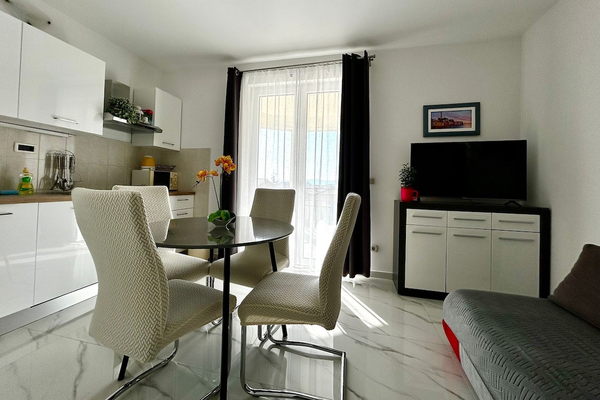 A modern kitchen with white cabinets, a dining table with chairs, a flat-screen TV on a stand, and a sofa in a living area.