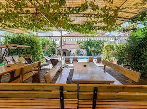 Eine Außenpatio mit Holzbänken und -tischen, ein Pool im Hintergrund und ein überdachter Bereich mit Dach.
